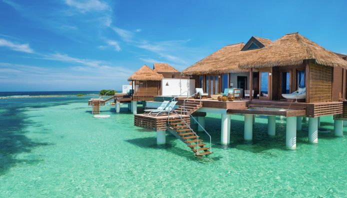 Thatched-roof overwater villas stand on stilts above clear turquoise water in the Maldives at one of the best all-inclusive resorts, with lounge chairs and steps leading directly into the sea, under a bright blue sky.