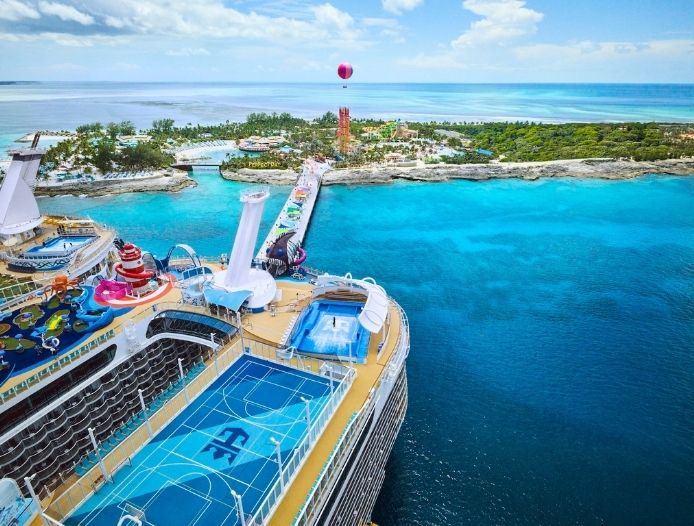 A large cruise ship with sports courts on its deck is docked near a tropical island, offering unforgettable cruise vacations amid clear blue water, lush greenery, and a tall red observation tower in the background.