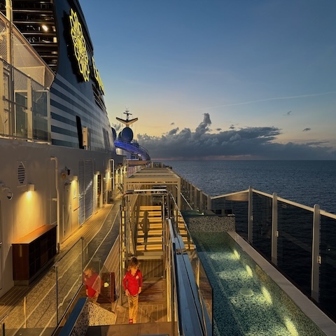 A grand staircase with glass railings descends to a stylish, modern lobby reminiscent of luxury cruise vacations, featuring patterned floors and seating beneath an ornate chandelier and circular ceiling lights.